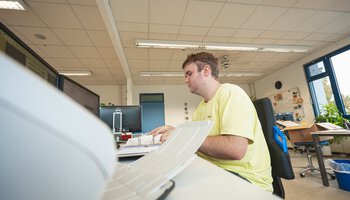Ein Mann sitzt an einem PC-Arbeitsplatz und sichtet Unterlagen | © Wendelstein Werkstätten