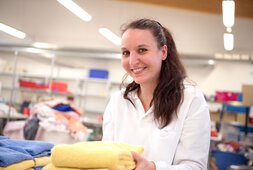 Eine Mitarbeiterin hält saubere und zusammengelegte Wäsche in der Hand und lächelt  | © Wendelstein Werkstätten